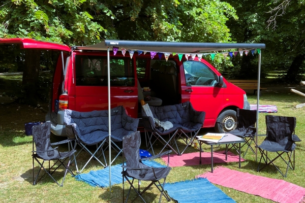 Hier sieht man ein Bild von unserem Streetwork Wohnzimmer. Zu sehen ist unser roter VW Bus mit ausgefahrener Markise, Campingstühle und Sofas im Stadtpark